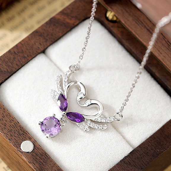 Silver heart-shaped pendant necklace with purple gemstones, displayed on a white cushion inside a wooden box over an open book.