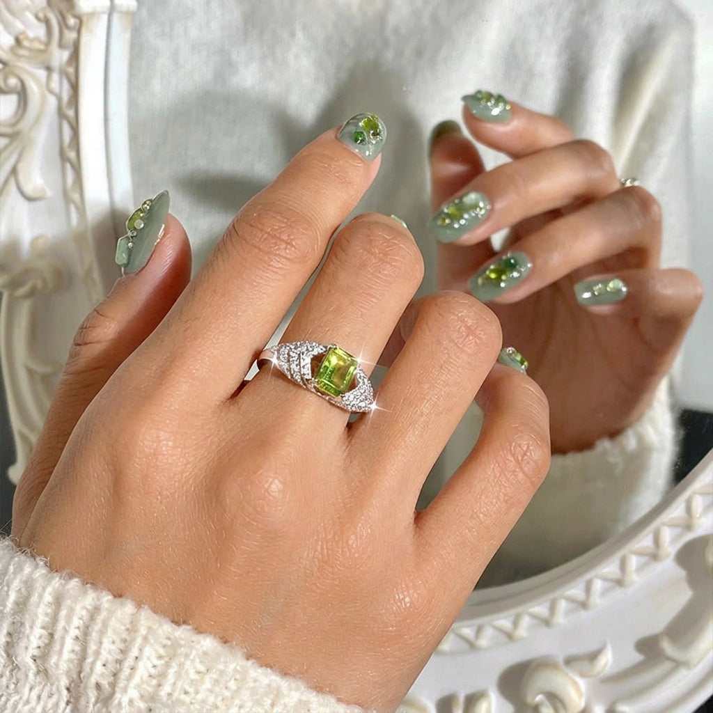 Hand wearing a ring with a green gemstone and diamond accents, against a neutral background.